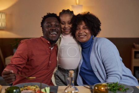 Foto de Cintura hacia arriba retrato de feliz familia negra con niña abrazando y sonriendo a la cámara en la mesa de la cena - Imagen libre de derechos