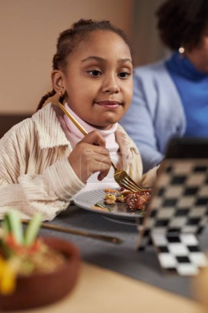 Téléchargez les photos : Portrait vertical de fille afro-américaine manger le dîner et regarder des vidéos en ligne via téléphone mobile - en image libre de droit