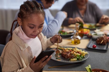 Porträt eines kleinen schwarzen Mädchens mit Smartphone am Esstisch, während es zu Hause mit der Familie isst