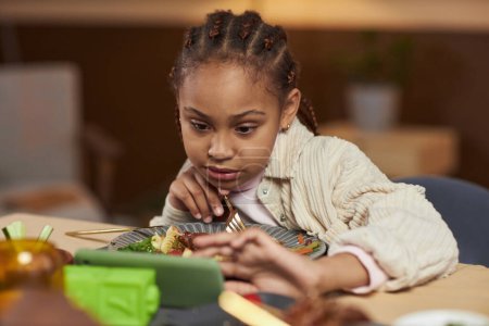 Photo for Front view of little African American girl using smartphone and watching videos while eating at dinner table - Royalty Free Image