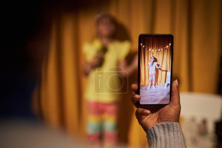 Foto de Primer plano de los padres sosteniendo el teléfono inteligente y filmando a una niña pequeña actuando en el show de talentos en casa con enfoque selectivo en la pantalla del teléfono - Imagen libre de derechos