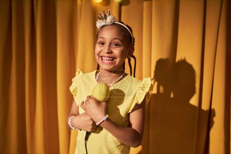 Photo for Waist up portrait of smiling little girl speaking to microphone while giving home concert against makeshift yellow curtains copy space - Royalty Free Image