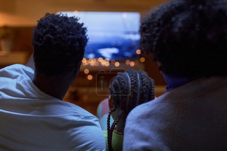 Photo for Back view of Black family with little girl watching Tv together in dark room sitting on sofa at home - Royalty Free Image