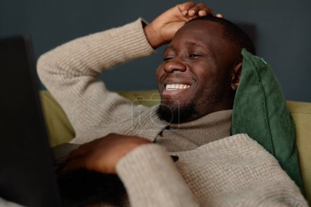 Téléchargez les photos : Portrait d'un homme afro-américain souriant allongé sur un canapé et regardant un écran d'ordinateur portable tout en se relaxant au travail - en image libre de droit