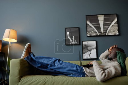 Téléchargez les photos : Vue latérale pleine longueur chez Black man allongé sur le canapé et regardant l'écran d'ordinateur portable tout en se relaxant au travail - en image libre de droit