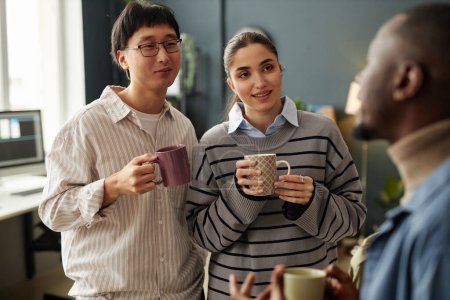 Photo for Candid waist up portrait of three adults chatting and holding mugs during coffee break in office - Royalty Free Image
