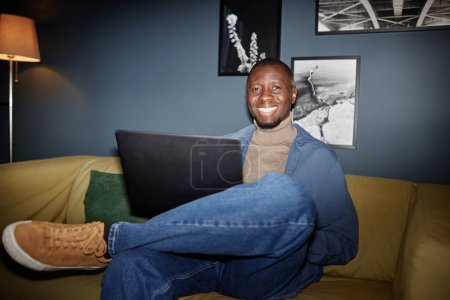 Foto de Retrato del hombre afroamericano adulto sonriendo a la cámara sentado en el sofá con el ordenador portátil, filmado con flash - Imagen libre de derechos