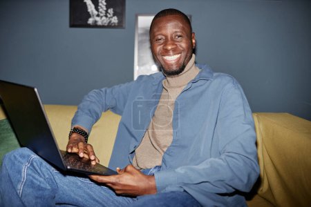 Téléchargez les photos : Portrait d'homme noir adulte joyeux assis sur le canapé avec un ordinateur portable et regardant l'appareil photo, tourné avec flash - en image libre de droit