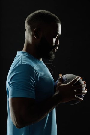 Foto de Retrato dramático de vista lateral del jugador de fútbol afroamericano sosteniendo la pelota contra fondo negro con contorno - Imagen libre de derechos