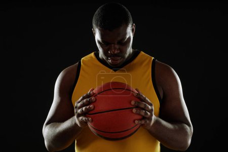 Téléchargez les photos : Portrait à la taille dramatique d'un joueur de basket-ball musclé tenant le ballon à la poitrine avec espace de copie contour contre-jour - en image libre de droit