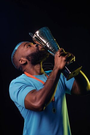 Téléchargez les photos : Portrait vertical d'un sportif afro-américain embrassant une coupe trophée et célébrant la victoire - en image libre de droit