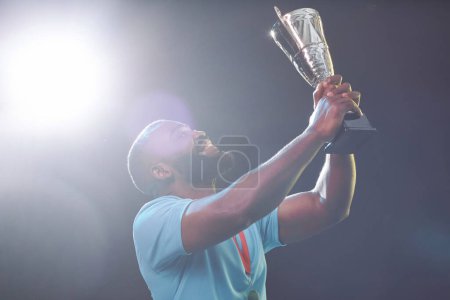 Téléchargez les photos : Portrait latéral d'un sportif afro-américain tenant une coupe trophée et applaudissant tout en célébrant la victoire avec un espace de copie de fusée éclairante - en image libre de droit