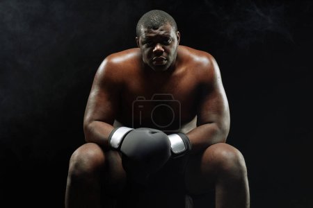 Foto de Dramático retrato retroiluminado del musculoso boxeador afroamericano mirando a la cámara sentada sobre fondo negro - Imagen libre de derechos