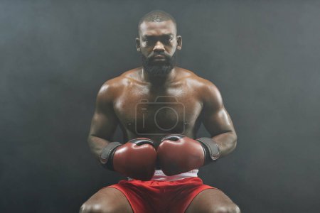 Foto de Retrato de vista frontal del duro boxeador afroamericano con guantes rojos y mirando a la cámara sentada sobre fondo negro - Imagen libre de derechos