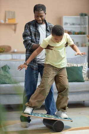 Foto de Retrato vertical de cuerpo entero del joven padre negro enseñando a su hijo de pie en el tablero de equilibrio y divertirse juntos - Imagen libre de derechos