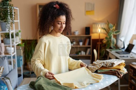 Waist up portrait of smiling young woman with big curly hair folding clothes on ironing board at home copy space