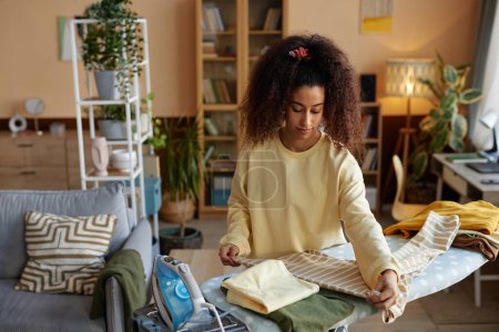 Photo for High angle portrait of curly haired young woman ironing and folding clothes on laundry day and taking care of house chores copy space - Royalty Free Image