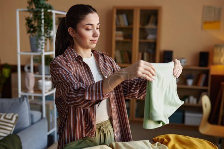 Waist up portrait of smiling young woman folding clothes on laundry day at home and enjoying chores copy space