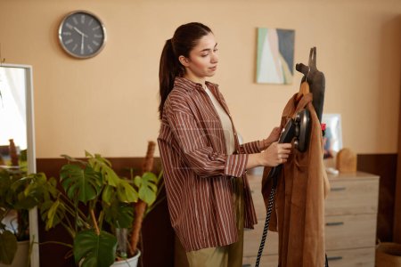 Photo for Side view portrait of young woman using garment steamer on velvet jacket in cozy home copy space - Royalty Free Image