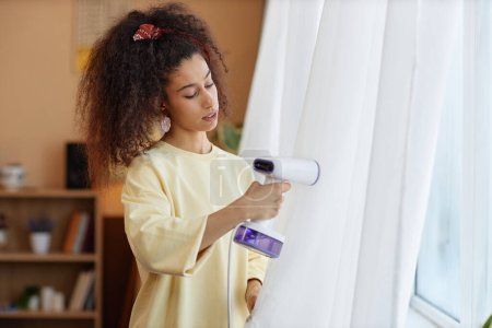 Waist up portrait of young woman steaming gauze curtains on window with with hand steamer during Spring cleaning copy space