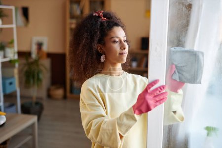 Porträt einer jungen Afroamerikanerin mit lockigem Haar, die zu Hause Fenster putzt und rosa Gummihandschuhe trägt
