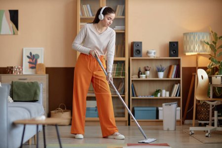 Photo for Full length portrait of young woman wearing headphones and mopping floors at home while enjoying cleaning copy space - Royalty Free Image