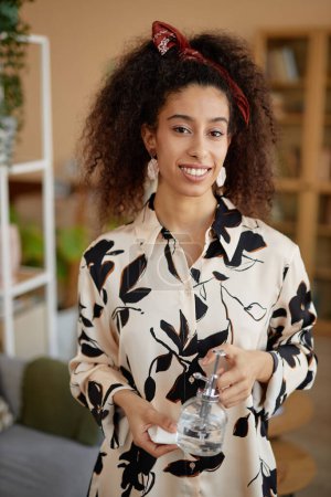 Téléchargez les photos : Portrait vertical de jeune femme noire aux cheveux bouclés tenant minuscule arrosoir et souriant à la caméra - en image libre de droit