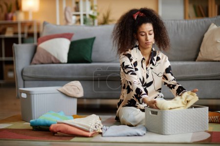 Téléchargez les photos : Portrait de jeune femme afro-américaine pliant et rangeant des vêtements propres tout en nettoyant la maison le week-end - en image libre de droit