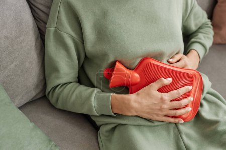 Foto de Primer plano de la mujer irreconocible sosteniendo botella de agua caliente roja al estómago que sufre de calambres menstruales espacio de copia - Imagen libre de derechos