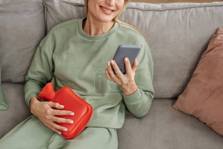 Photo for Close up of smiling woman holding red hot water bottle to stomach and using smartphone at home copy space - Royalty Free Image