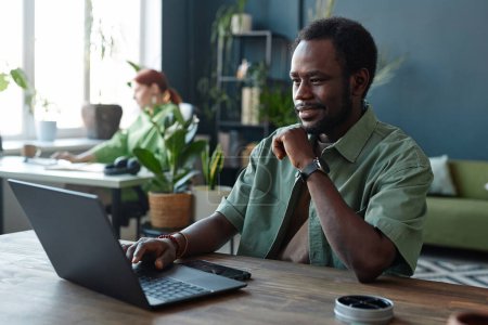 Foto de Retrato de un hombre afroamericano sonriente usando una computadora portátil en el lugar de trabajo en una oficina abierta decorada con plantas verdes - Imagen libre de derechos