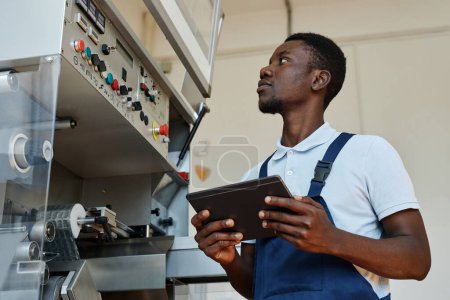 Vista lateral del trabajador de fábrica afroamericano sosteniendo la tableta mientras opera unidades de la máquina y supervisa la producción, espacio de copia