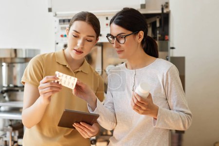 Retrato de cintura hacia arriba de dos mujeres jóvenes mirando las píldoras de embalaje supervisando las producciones en la fábrica farmacéutica