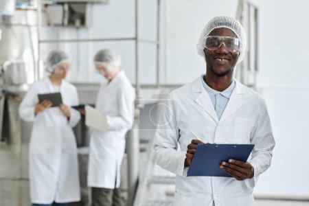 Photo for Waist up portrait of black young man wearing lab coat and smiling at camera in clean workshop of pharmaceutical factory, copy space - Royalty Free Image