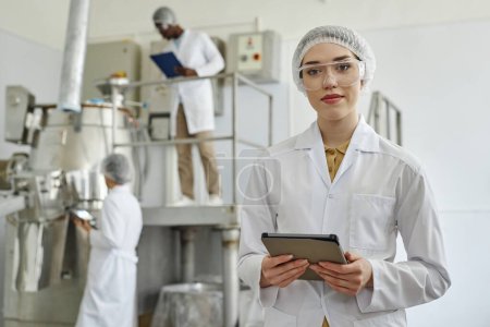 Waist up portrait of modern young woman wearing lab coat and looking at camera in clean workshop of pharmaceutical factory, copy space