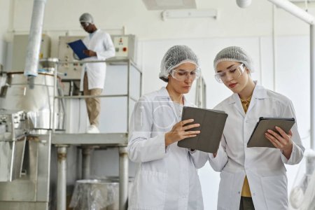 Retrato de cintura hacia arriba de dos mujeres jóvenes con batas de laboratorio y uso de tableta digital en el taller de fábrica farmacéutica, espacio de copia