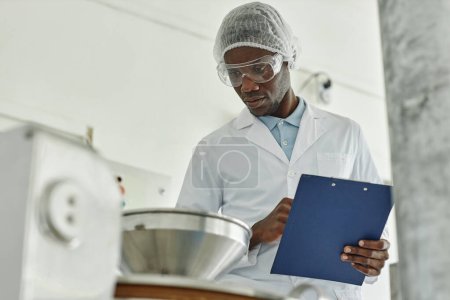 Retrato del joven negro usando bata de laboratorio y sujetando el portapapeles mientras supervisa la producción en la fábrica de alimentos, espacio para copiar
