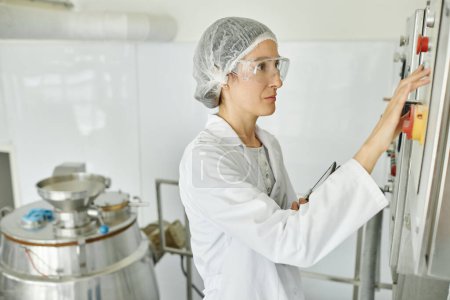 Retrato de vista lateral de mujer adulta que usa bata de laboratorio mientras opera el equipo en el taller de fábrica y presiona los botones en el panel de control, espacio de copia