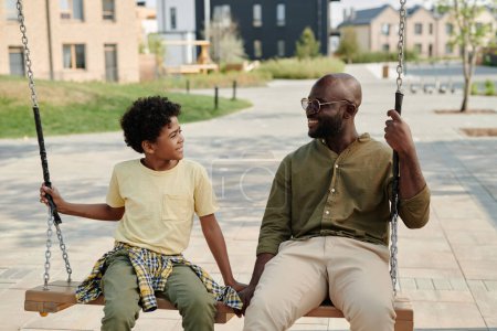 Téléchargez les photos : Afro-américain papa balançant sur swing avec son fils pendant leur promenade à l'extérieur - en image libre de droit