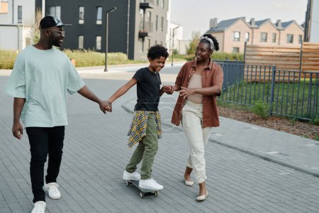 Foto de Familia feliz caminando por la calle tomados de la mano, le enseñan a su hijo a montar en monopatín al aire libre - Imagen libre de derechos