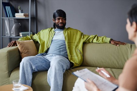 Téléchargez les photos : Afro-Américain assis sur le canapé et discutant de ses problèmes avec un psychologue au bureau - en image libre de droit