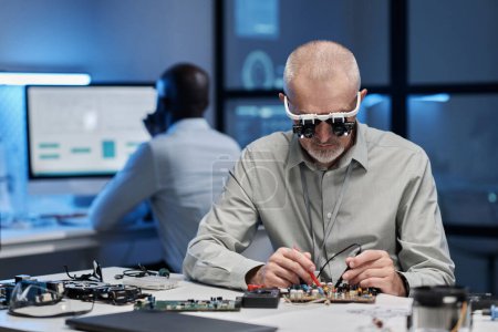 Foto de Ingeniero maduro en gafas especiales concentrándose en su trabajo con chip de computadora en la mesa en el laboratorio - Imagen libre de derechos