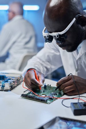Téléchargez les photos : Image verticale d'un ingénieur afro-américain utilisant des lunettes spéciales pour souder une carte mère pour un nouveau système informatique - en image libre de droit