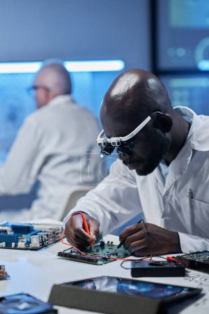 Téléchargez les photos : Image verticale d'un ingénieur afro-américain travaillant avec une carte mère d'ordinateur à table au bureau - en image libre de droit