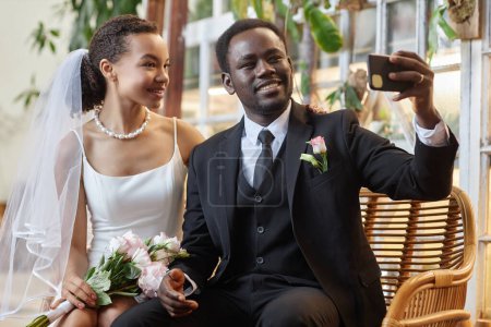 Photo for Portrait of young black couple as bride and groom taking selfie photo together in green orangerie - Royalty Free Image