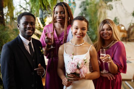 Téléchargez les photos : Portrait taille haute de jeune couple afro-américain comme mariée et marié posant avec des demoiselles d'honneur lors de la cérémonie de mariage et souriant à la caméra - en image libre de droit