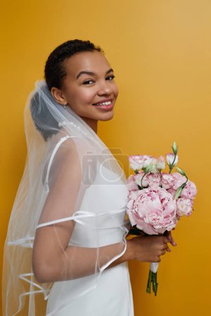 Téléchargez les photos : Portrait vertical de jeune femme noire portant une robe de mariée et tenant des fleurs sur fond jaune - en image libre de droit
