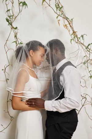 Photo for Vertical portrait of elegant black couple as bride and groom embracing affectionately standing against wall decorated with plants and vines - Royalty Free Image