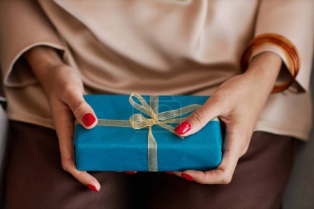 Foto de Manos de mujer joven en blusa de seda beige sosteniendo una pequeña caja de regalo azul con regalo de cumpleaños mientras está sentada frente a la cámara en la fiesta - Imagen libre de derechos