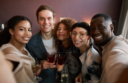 Photo for Happy young multicultural friends with glasses of wine and champagne looking at smartphone camera while posing for selfie at home party - Royalty Free Image